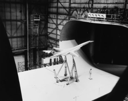 A scale model of an aircraft is being tested in a wind tunnel with a technician standing nearby in a large industrial facility, while CFD simulations complement the observations.