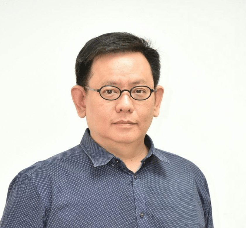 A man with short black hair, wearing glasses and a blue button-up shirt, stands against a plain white background.