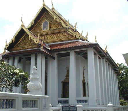 A traditional Thai temple with ornate golden roof decorations and white pillars, surrounded by a white fence and greenery.