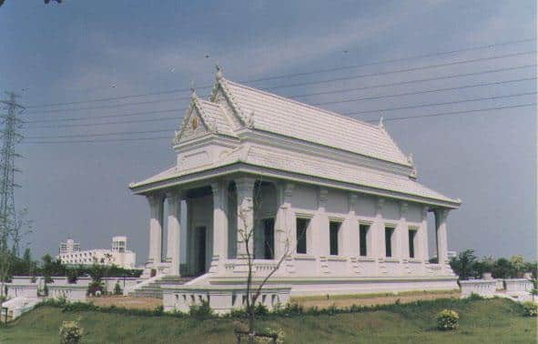 A white temple with a traditional, ornate roof stands on a grassy area under a cloudy sky, with power lines and a building in the background.