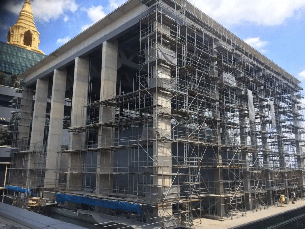 A large building is covered in scaffolding, indicating construction or renovation work. A golden spire is visible in the background against a partly cloudy sky.