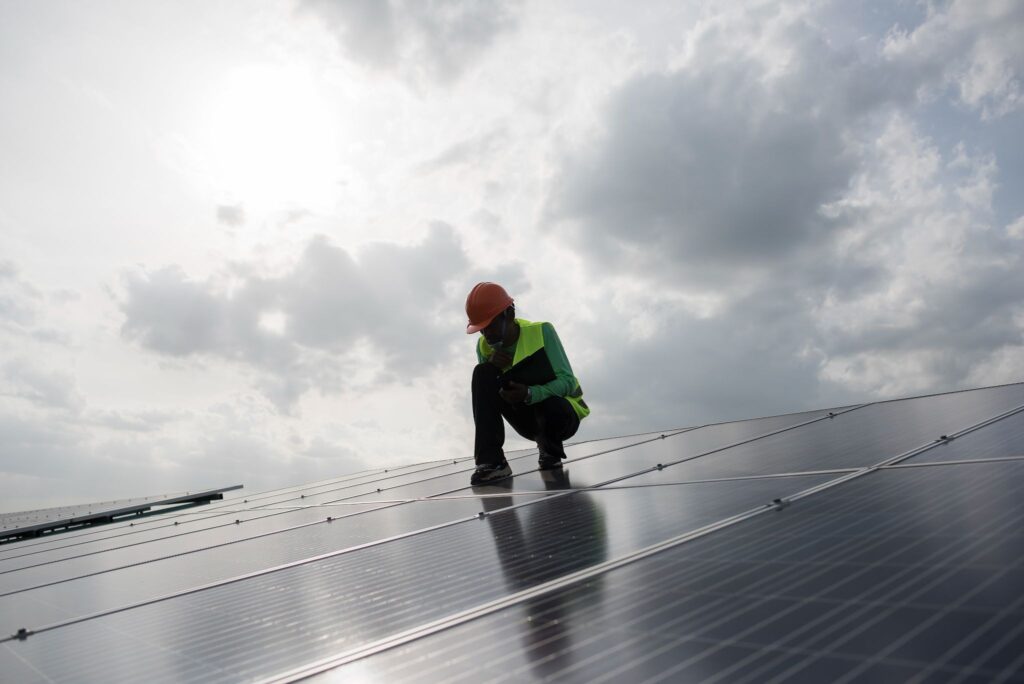 A worker in a hard hat and safety vest inspects solar panels on a rooftop under a cloudy sky with sunlight filtering through.